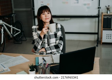 successful asian businesswoman is smiling and looking into the distance while enjoying tea and refreshing morning at desk in the office with daylight. - Shutterstock ID 2176965341