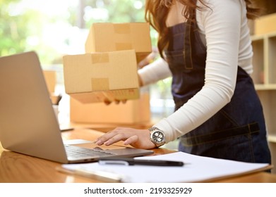 Successful Asian Female Online Business Owner Or SME Entrepreneur Preparing Shipping Packages And Update Packing Status On Her Website In Laptop Computer. Cropped Image