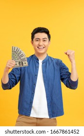 Successful Asian Man. Happy Young Man Holding Money While Standing And Arms Up, Isolated On Yellow Background