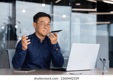 Successful asian man inside office working with laptop, businessman recording audio message using app on smartphone, man using artificial intelligence assistant to help finding a solution. - Powered by Shutterstock