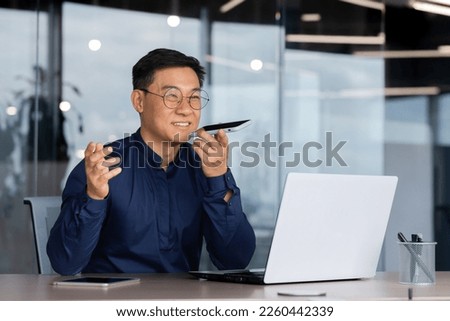 Successful asian man inside office working with laptop, businessman recording audio message using app on smartphone, man using artificial intelligence assistant to help finding a solution.