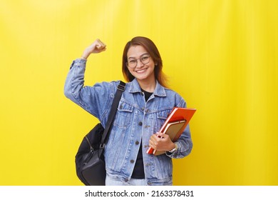 Successful asian young woman standing while clenching hand and holding books. Isolated on yellow