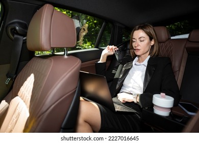 Successful attractive businesswoman sitting in the passenger seat in her luxury car working on laptop and driving to a business meeting