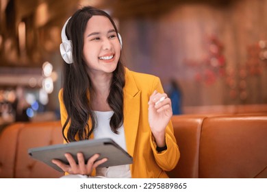 successful attractive smart business casual Asian female woman using tablet device working online remotely in cafe mall while listening music from headphone carefree working on holiday weekend - Powered by Shutterstock