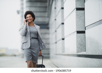 A successful black business woman walking with her luggage and using smart phone outside of the airport.