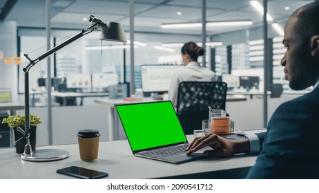 Successful Black Businessman Sitting at Desk Working on Green Screen Chroma Key Laptop Computer in Big City Office. Hard Working Top Manager Doing Research for e-Commerce Project. Over Shoulder - Powered by Shutterstock