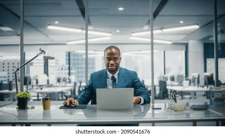 Successful Black Businessman In Tailored Suit Working On Laptop Computer On Top Floor Office Overlooking Big City. Professional CEO Managing Environmental, Social And Corporate Governance