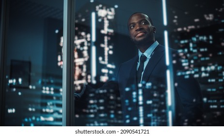 Successful Black Businessman In A Tailored Suit Standing In His Office Looking Out Of The Window On Night City. Successful Investment Manager Working Late Planning E-Commerce Service Purchase Strategy