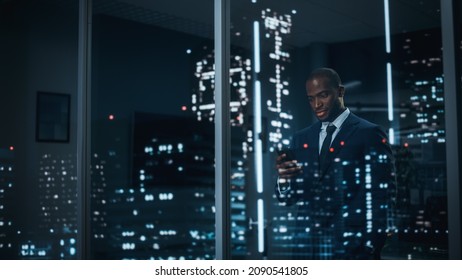 Successful Black Businessman In A Tailored Suit Standing In His Office Looking Out Of The Window On Night City. Successful Investment Manager Working Late. Stylish Outside Shot