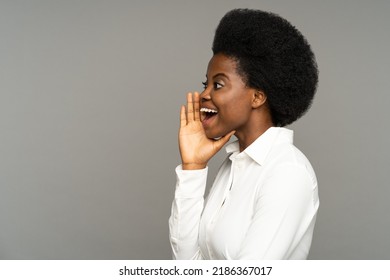 Successful Black Businesswoman With Kinky Hair Holds Hand Near Mouth Speaking. Young African American Woman In Classic White Shirt Says Advertising Text In Empty Space Standing On Grey Background