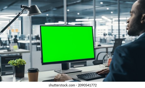 Successful Black Businesswoman Sitting at Desk Working on Green Screen Laptop Computer in Office. African American Businessperson using Chroma Key Display. Over Shoulder Shot