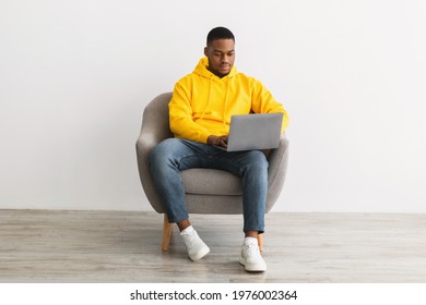 Successful Black Freelancer Guy Using Laptop Working Online Sitting In Armchair On Gray Wall Background Indoors. African American Millennial Man Browsing Internet On Computer Writing Article For Blog