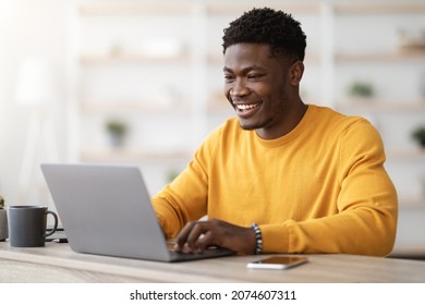 Successful black guy project manager sitting at workdesk at office, typing on laptop keyboard and smiling, chatting with clients, enjoying his job, copy space. Freelancer working from home