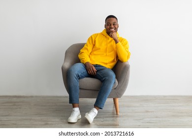 Successful Black Millennial Guy Smiling To Camera Touching Chin Sitting In Armchair Near Gray Wall Indoors, Wearing Yellow Hoodie. African American Man Relaxing Posing At Home. Full Length - Powered by Shutterstock