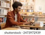 Successful black student guy sitting at desk in university library, with laptop, doing homework or preparing for exam, browsing internet, free space