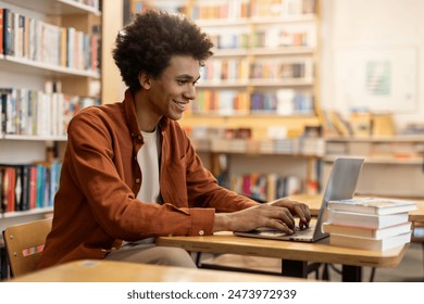 Successful black student guy sitting at desk in university library, with laptop, doing homework or preparing for exam, browsing internet, free space