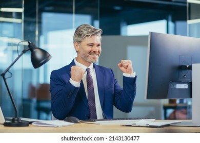 Successful Boss Works In A Modern Office At The Computer, A Senior Man Gray And Happy With A Beard Looks At The Camera And Smiles, Holds His Hands Up, Celebrates Victory