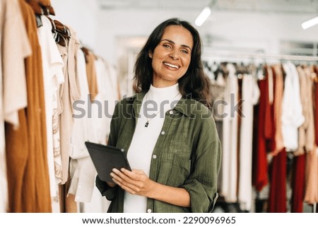 Successful boutique owner is standing in her store, her tablet in hand, as she manages her business operations with ease. Happy business woman taking inventory for her small business.