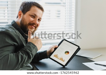 Successful broker showing thums up and using tablet with graphs and diagrams on screen. Concept of enjoying successs and business project. Gladden man working with modern gadget at office.