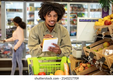 Successful Budget Planning. Happy African American Man Writing Grocery Shopping List Calculating Food Prices Standing With Cart In Supermarket. Financial Literacy, Household Economy Concept