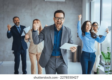 Successful business group celebrating an achievement at the office with arms up and looking at the camera smiling. Group of young businesspeople huddled together in solidarity in a modern office - Powered by Shutterstock