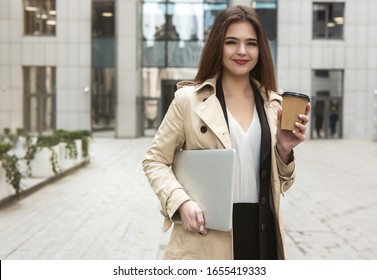 Successful Business Lady Holding Laptop Under Arm Walking Down The Street Near Office Center With Cup Of Coffee Looking Pleased, Eating On The Go Concept