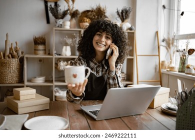 Successful business lady owner of shop made of clay talking to customer. Caucasian female potter entrepreneur talking to buyer of handmade mug sitting at table and taking order using laptop. - Powered by Shutterstock