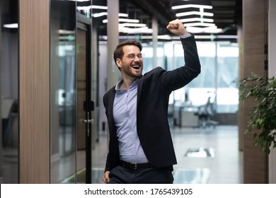 Successful Business Man Celebrating Victory With Raised Fist. Happy Millennial Businessman Cheering To Success In Office Corridor. Excited Smiling Employee Getting Dream Job Or Boss Conducting Deal