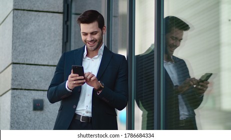 Successful Business Man Standing In Black Suit At Street. Young Businessman Using Smartphone Outside. Professional Businessman Smiling With Mobile Phone At Street.