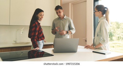 Successful Business People Negotiate With Clients At Online Conference At Lockdown. Three Young People Standing At Modern Kitchen While Talking With Clients At Laptop. Working From Home Concept, Wide