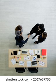 Successful Business People Shaking Hands In The Modern Office. Laptop Computer, Tablet, Mobile Phone, Coffee Cup And Document Are On The Table. Top View