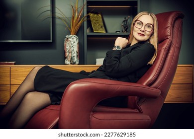 Successful business woman in elegant black dress and glasses. A beautiful stylish middle-aged woman sits in an expensive leather chair in a modern office and smiles. 