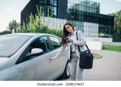 Successful business woman standing outside the office building while entering the car on a driver seat and using a smartphone. Businesswoman wearing a suit and a business bag. Woman leader. - Powered by Shutterstock