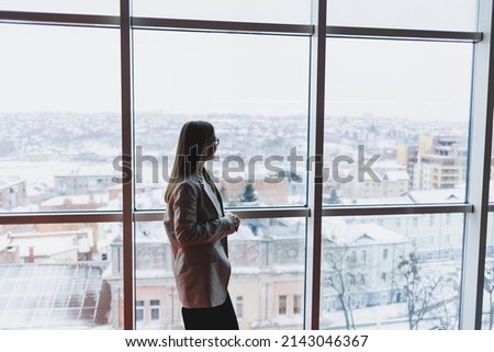 A successful business woman in trousers and a jacket stands and looks out the huge panoramic window of the business center skyscraper.