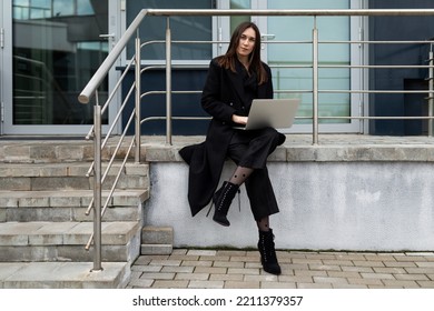 successful business woman working on laptop online sitting on stairs wearing stylish black coat