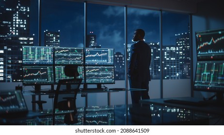 Successful Businessman Looking Out of the Window on Late Evening. Modern Hedge Fund Office with Computer with Multi-Monitor Workstation with Real-Time Stocks, Commodities and Exchange Market Charts.