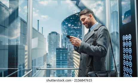 Successful Businessman Riding Glass Elevator To Office In Modern Business Center. Handsome Happy Man Smile While Using Smartphone, Write Text Message, Check Social Media And Work Emails In A Lift.