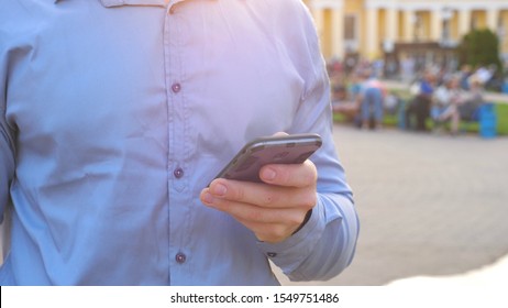 Successful Businessman In Shirt Standing Among City Street And Browsing Smartphone. Confident Man Typing Text Or Using App On His Phone During Break In Work. Blurred Background. Slow Mo Dolly Shot