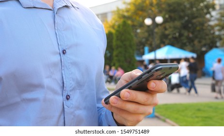 Successful Businessman In Shirt Standing Among City Street And Browsing Smartphone. Confident Man Typing Text Or Using App On His Phone During Break In Work. Blurred Background. Slow Mo Dolly Shot