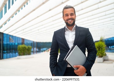 Successful Businessman In Suit With Beard Standing In Front Of Office Building Confidently Looking At Camera And Smile Holding Document. Hispanic Male Business Person.