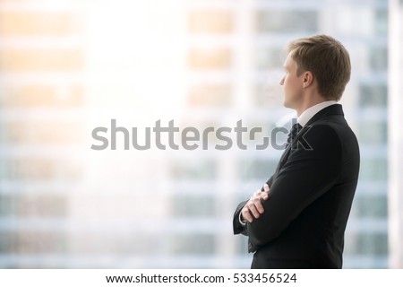Successful businessman in suit standing in office with hands crossed on chest, looking through window at big city buildings, planning new projects, waiting for meeting to start. Copy space for text
