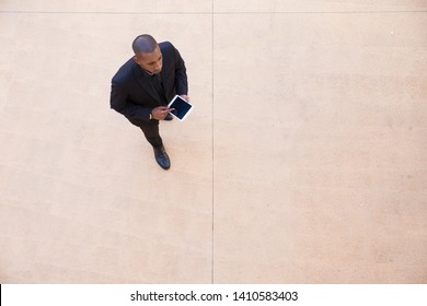 Successful Businessman With Tablet Walking Through Office Lobby. Top View Of Black Man In Formal Suit Using Tablet And Walking. Communication Concept