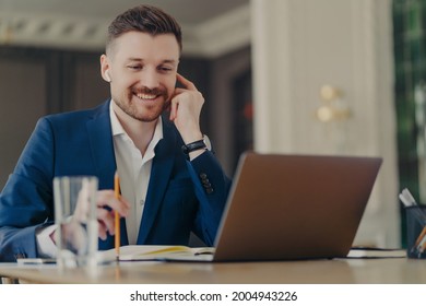 Successful Businessman In Wireless Earphones Holding Web Conference At His Office With Employees, Wearing Formal Dark Blue Suit While Sitting In Front Of Laptop With Pencil In Hand, Working At Home