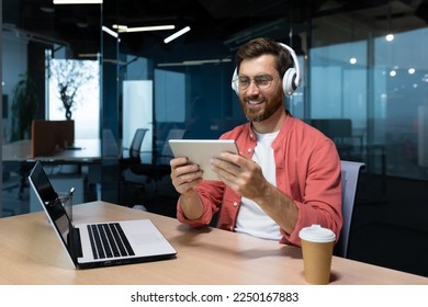 Successful businessman at workplace smiling man watching online video sitting at desk wearing headphones and glasses programmer holding tablet computer inside office.