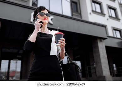 Successful businesswoman leader stands in big city talks on mobile phone. Smiling woman making business call on cell walking with cup of coffee on background of office centre, multitasking outdoors.