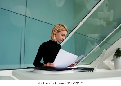 Successful Businesswoman Reviewing Monthly Reports While Sitting With Portable Net-book In Office Interior, Young Female Intelligent Bookkeeper Reading Paper Documents During Work On Laptop Computer