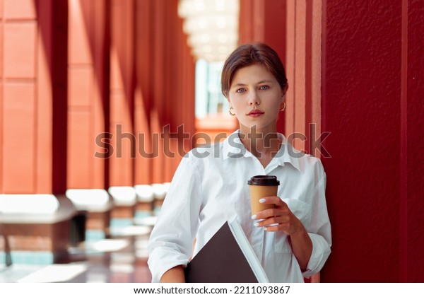 Successful businesswoman standing in central\
business district in modern city. Caucasian female business person\
with documents paper and coffee take away in white shirt on city\
street. Business