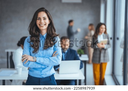Successful businesswoman standing in creative office and looking at camera. Young woman entrepreneur in a coworking space smiling. Portrait of beautiful business woman standing in front of team