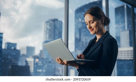 Successful Businesswoman In Stylish Dress Working On Laptop, Standing Next To Window In Big City. Confident Female CEO Analyze Financial Projects. Manager At Work Planning Marketing Campaign.