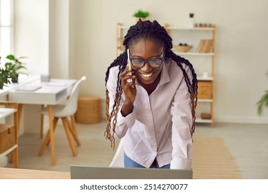 Successful businesswoman talks on phone. Happy beautiful young African American business woman in glasses with Afro hair standing by desk at home office, uses laptop computer and talks on mobile phone - Powered by Shutterstock